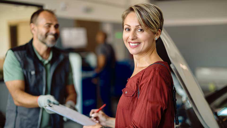 Frau hält einen Stift und unterschreibt Unterlagen in einer Werkstatt, im Hintergrund steht ein Mann